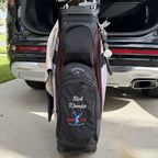 A black Callaway golf bag with red piping and custom white and blue embroidery, standing in front of an open black SUV trunk on a concrete driveway.