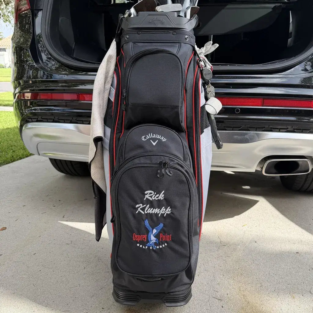 A black Callaway golf bag with red piping and custom white and blue embroidery, standing in front of an open black SUV trunk on a concrete driveway.