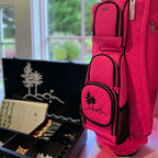 A bright pink quilted golf bag with black trim stands next to an open black presentation case containing golf accessories, photographed indoors near a window.