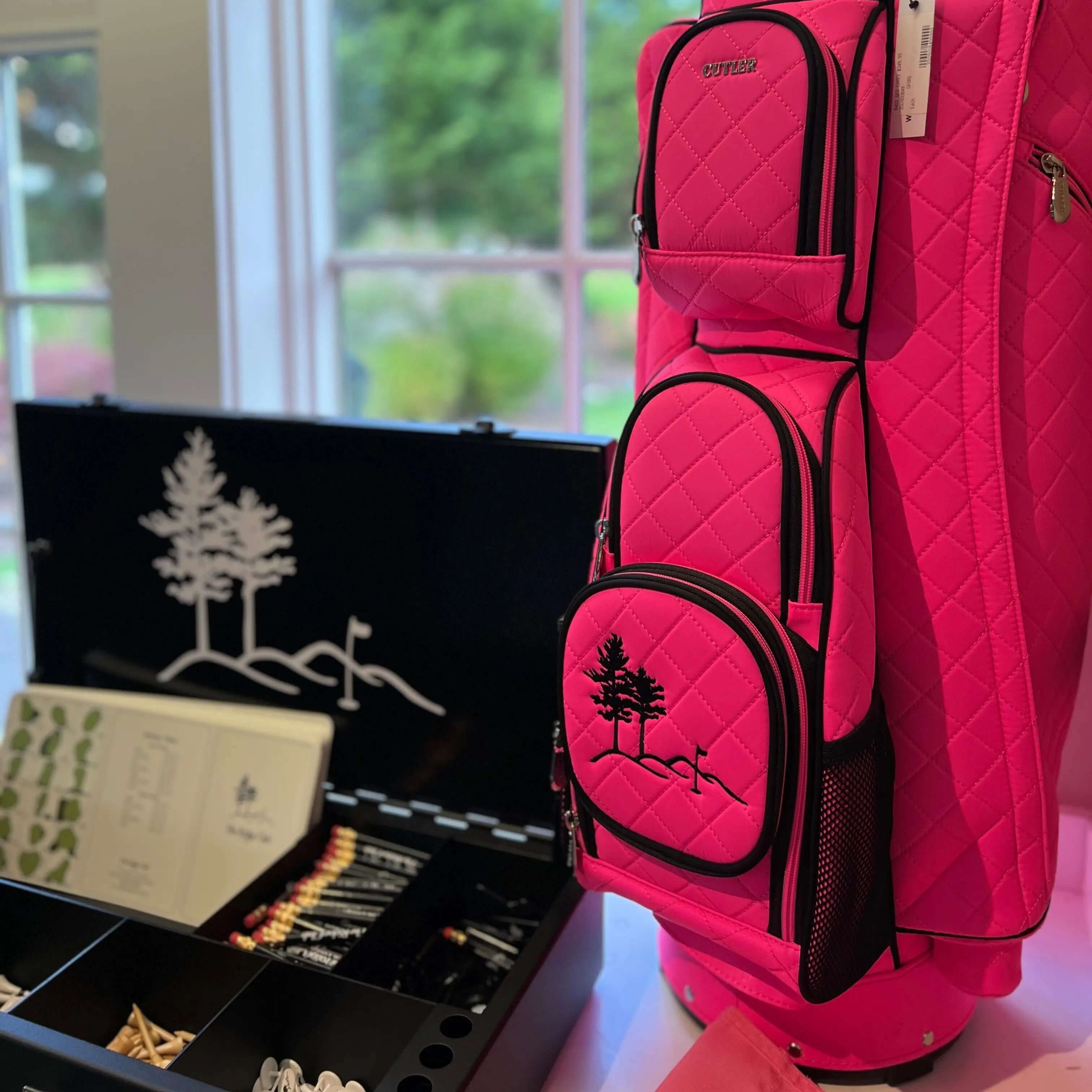 A bright pink quilted golf bag with black trim stands next to an open black presentation case containing golf accessories, photographed indoors near a window.