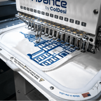 A close-up of an embroidery machine with a white fabric hoop containing a blue and white golf bag graphic, under large text reading 'CUSTOM GOLF BAG'. The machine branding 'Avance by ColDesi' is visible on the front.