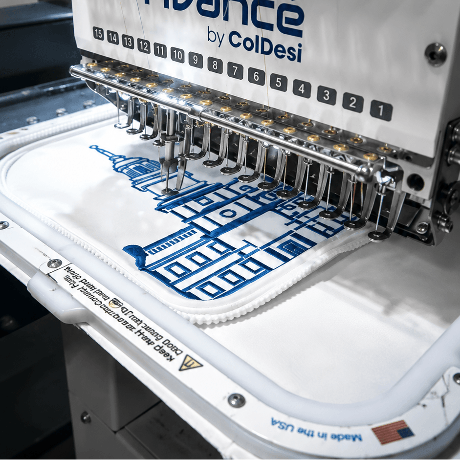 A close-up of an embroidery machine with a white fabric hoop containing a blue and white golf bag graphic, under large text reading 'CUSTOM GOLF BAG'. The machine branding 'Avance by ColDesi' is visible on the front.